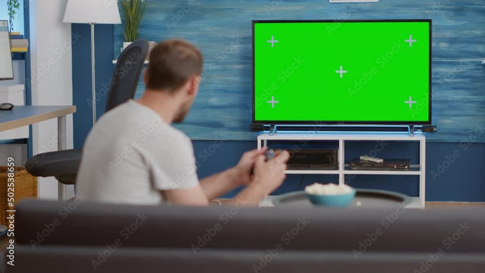 Poster man holding wireless controller playing console video game on green screen tv while sitting on sofa 
