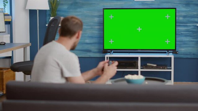 Man holding wireless controller playing console video game on green screen tv while sitting on sofa in modern living room. Gamer relaxing on couch enjoying online gaming on croma key display.