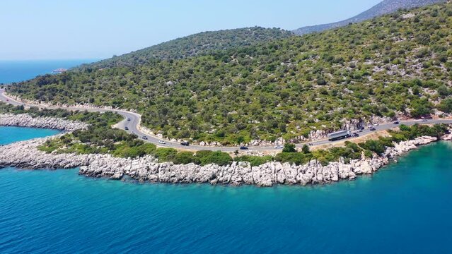 Wide drone aerial view of a coastal highway road near Finike in Turkey on a sunny summer day as cars and trucks drive by a large green hill