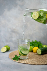 Pouring tasty fresh cucumber water from bottle into glass. A bottle of clean, cool and fresh drink stands on a gray concrete background with ingredients on board. Strengthening immunity, diet.