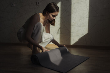 Side view full body of slim barefoot woman in white clothes rolling yoga mat on floor after...