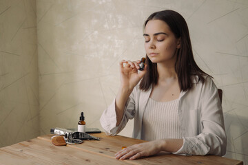 Relaxed woman with closed eyes smelling aromatic fragrance of natural essential oil from glass bottle while enjoying aromatherapy at table at home  - obrazy, fototapety, plakaty