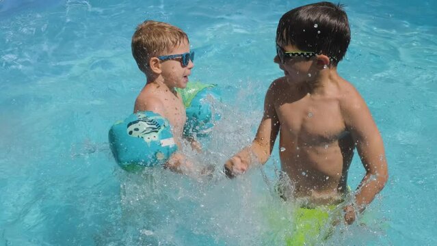 Happy boys in sunglasses waving arms and jumping in swimming pool water, Slow motion shot