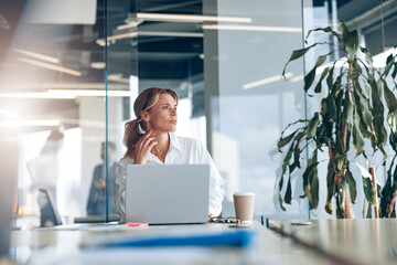 Focused businesswoman working on laptop and looking out the window at her workplace at modern office