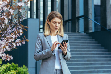 Fototapeta premium Serious and pensive business woman uses the phone outside the office, looking at the smartphone screen