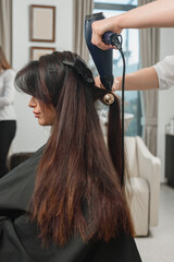 Young professional hairdresser drying long client's hair with blow-dryer in beauty salon
