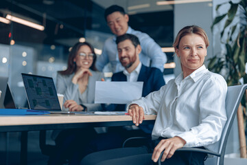 Reflective confident businesswoman sitting on meeting in office with her colleagues at background . 