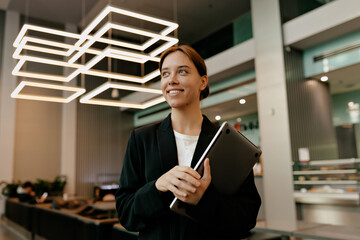 Smiling attractive female worker is holding laptop and looking aside with happy emotions in modern...