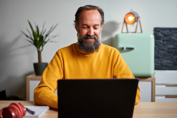 portrait of mature bearded man in yellow sweatshirt sitting at desk with laptop working on remote...