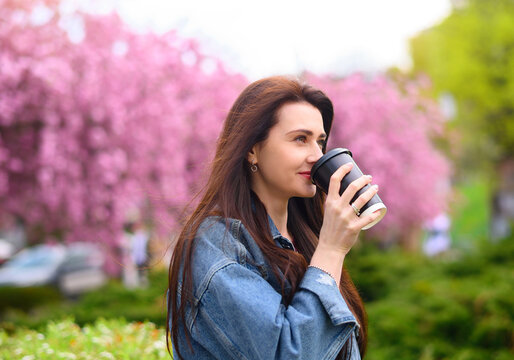 Young beautiful girl in a blue denim jacket drinks coffee on a background of flowering sakura rose tree. High quality photo