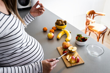 Smiling pregnant woman preparing fruit salad at home in kitchen.