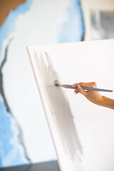 Woman's hand is painting big grey picture stands on the easel at home. Focus is at the brush. Art concept.