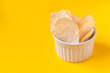 Riffled potato chips in white ceramic bowl on yellow background. Junk unhealthy food, chips, party food, fast food concept, free space for text