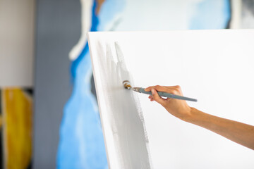 Woman's hand is painting big grey picture stands on the easel at home. Focus is at the brush. Art concept.