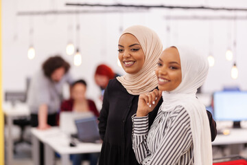 Two African American muslim female colleagues with hijab in a modern office space. Group of people working in the background Diversity and success concept..