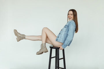 Side view of young smiling woman wearing denim jacket, beige boots, sitting on high stool, raising and stretching leg.