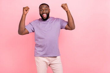 Photo of funny lucky young guy wear purple t-shirt rising fists empty space isolated pink color background