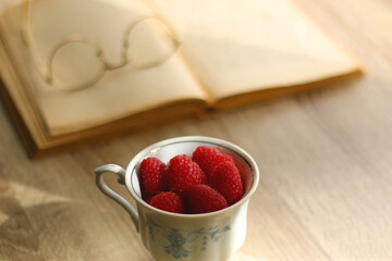 Porcelain cup filled with raspberries, open book, reading glasses and vase with flowers on the table. Selective focus.