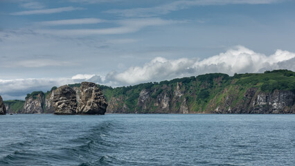 The picturesque coast of Kamchatka against the background of blue sky and clouds. There is green vegetation on the coastal hills. Sheer cliffs rise above the water. Avacha Bay.
