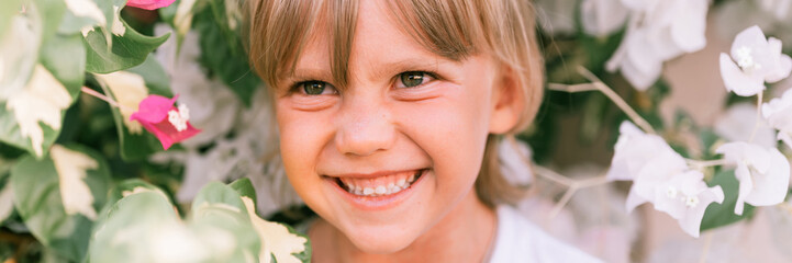 portrait of face candid little happy smiling five year old blonde kid boy with green eyes in pink and white flower plants in nature. children have fun summer holidays. bright light and airy. banner