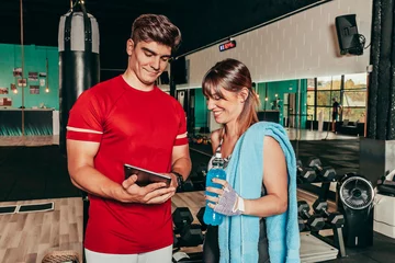 Foto op Canvas Smiling personal trainer showing woman results of training on tablet. Woman standing next to him and looking at tablet. Gym interior. People, fitness, sport and modern technology concept. © carlesmiro