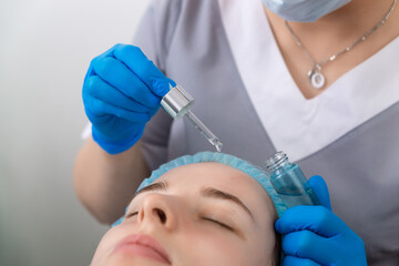 Hands of cosmetologist dripping nourishing oil facial skin of young woman in spa salon.