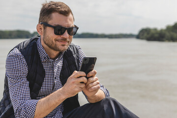 Mid adult man wearing casual clothes and sunglasses typing message on his smartphone while sitting...