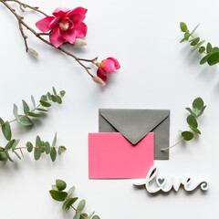 Pink magnolia flowers on twigs, fresh eucalyptus leaves. Blank pink card with grey envelope. Wood word love. Copy-space, place for greeting text. Chinese new year. Top view, off white background.