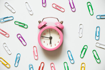 A pink alarm clock around multicolored paper clips lies on a white background, time