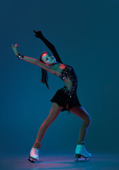 Dynamic portrait of young girl, female figure skater in black stage dress skating isolated on blue background in neon light. Concept of sport, beauty, active lifestyle.