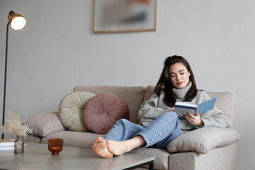 Young beautiful woman wearing grey turtleneck reading a book at home. Brunette female in a sweater...