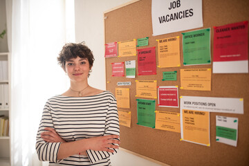 Happy young woman student standing in front of employment noticeboard.