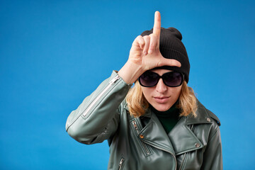 Portrait of young woman with blonde hair in green jacket standing, showing loser gesture with hand...