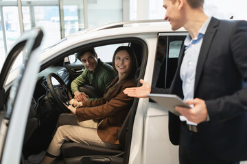 Fototapeta na wymiar Millennial car salesman discussing purchase of new auto with happy young couple after test drive at dealership centre
