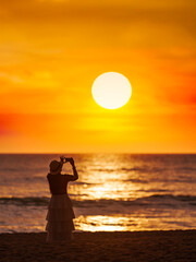 Fototapeta na wymiar Young women come to travel to relax at the sea on vacation using A smartphone takes pictures of the sunset silhouette alone in the evening.