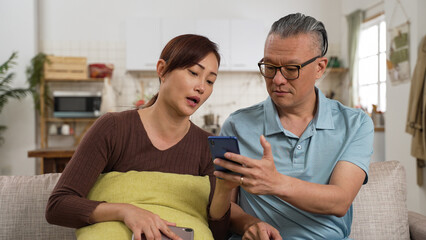 asian adult daughter helping senior dad with problem while they are using smartphones together in living room at home. she explains and shows him how to use
