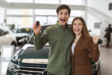 Overjoyed young Caucasian couple making YES gesture, showing car key, buying new auto at...