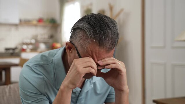 Worried older unhealthy asian man sitting on the couch. Unhappy frustrated elderly grandfather listening to bad news in the bright living room at home.