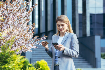 Fototapeta premium Beautiful blonde woman near a tree with flowers holds a phone and a bank credit card, smiles and makes purchases in the online store