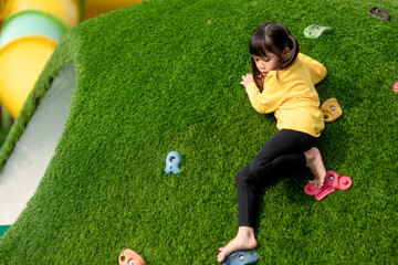 Cute Asian girl having fun trying to climb on artificial boulders at schoolyard playground, Little...