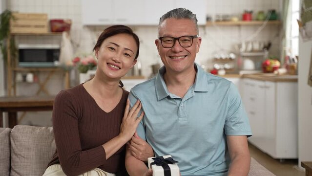 portrait of asian retired father and adult daughter smile and looking at camera with Father’s Day gift in living room at home. the smiling woman hugs the man and leans on his shoulder