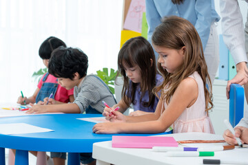Fototapeta premium Selective focus of a little cute Caucasian girl sitting at a small table, drawing intentionally together with multi-ethnic friends, with teachers standing watching them in the back at a kindergarten.