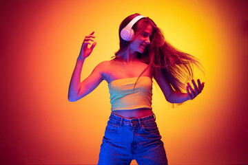 Portrait of young emotive girl listening to music in headphones and dancing isolated over gradient...