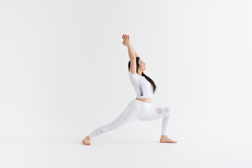 a beautiful young girl with dark hair stands in the pose of Virabhadrasana 1 on a white background. Yoga class