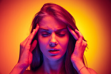 Close-up portrait of young girl touching temples, suffering from headache isolated over gradient red yellow background in neon