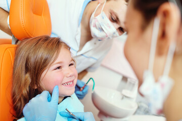 young dentists, a man and a woman, examine the teeth of a child's patient - a little pretty girl who is sitting in an orange dental chair.