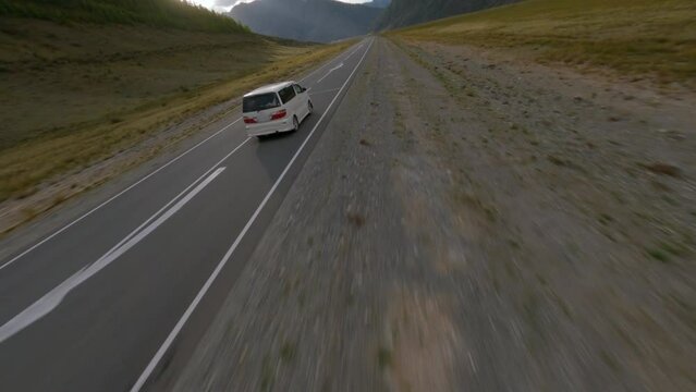 FPV sport drone shooting white car minibus riding highway from shadow sunlight adventure journey transportation picturesque mountain scenery. Aerial view minivan driving on road at cliff valley park