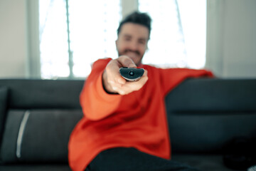 Young man wearing red hoodie holding television remote control with big smiles on sofa. Watching TV while sitting on the couch at home. Selective focus on remote tv controller.