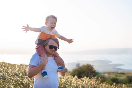 Smiling Kid Blond Boy Sitting On Dad Shoulders In Nature On A Windy And Sunny Day