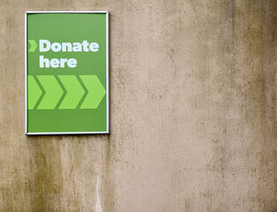 Green sign saying "Donate here" against rough worn out concrete wall. Off-center so lots of room for copy text.